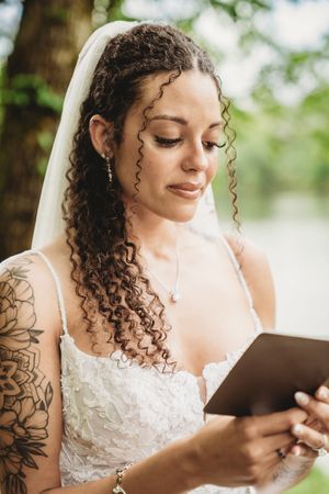 Bride with curly hair and tattoos reads vows from a book outdoors, wearing a white lace dress and veil.