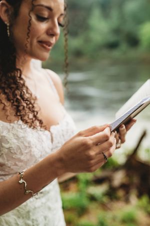 A bride in a white lace dress reads her wedding vows from a book outdoors near a river.