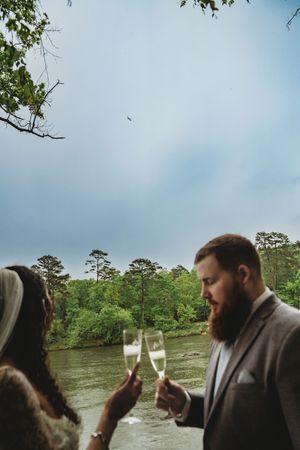 Bride and groom toasting with champagne flutes by a river, surrounded by lush green trees on their wedding day.