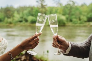 Bride and groom toasting with engraved Mr. and Mrs. champagne flutes near a scenic river backdrop.