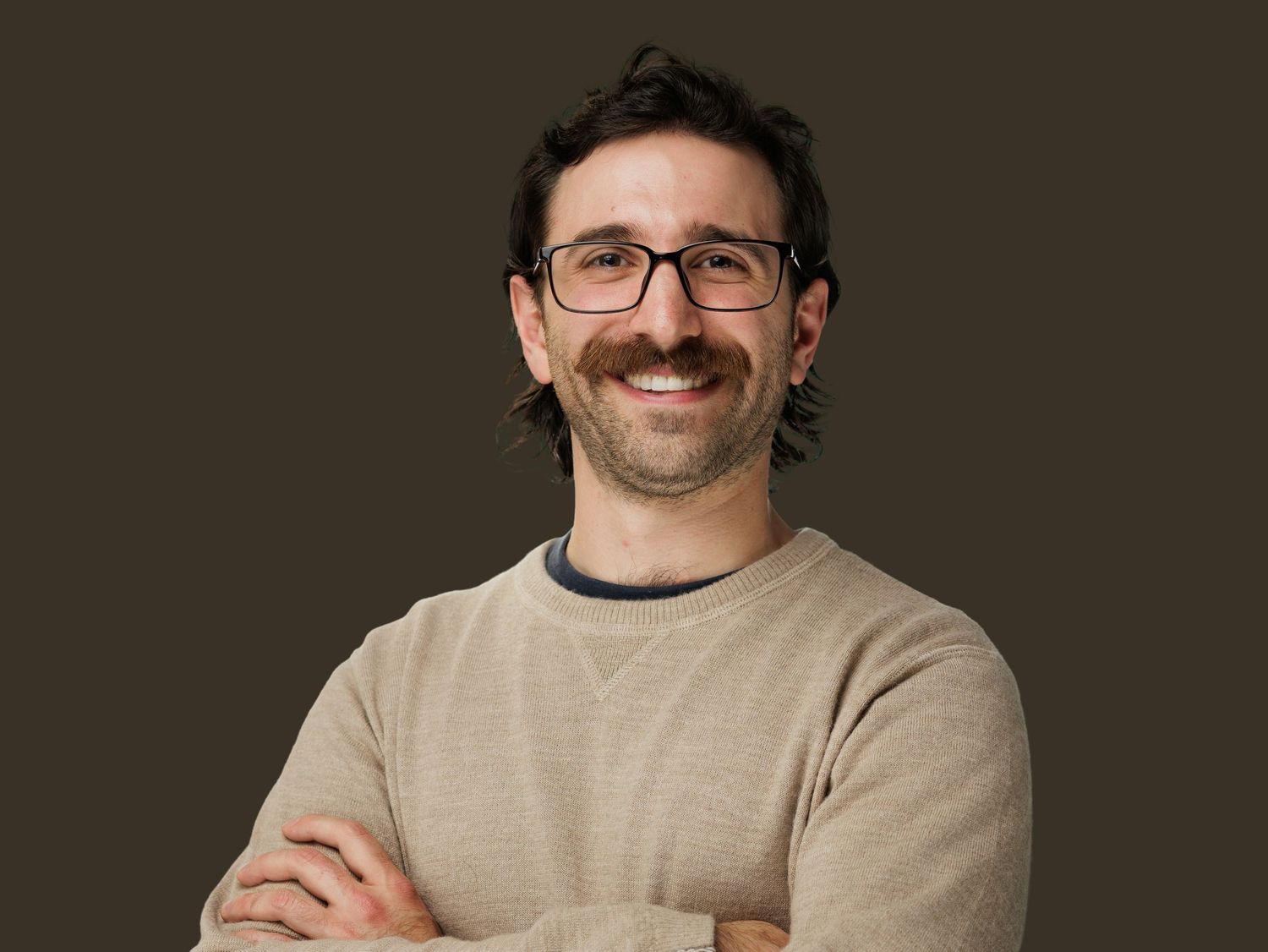 Smiling man with glasses and beard wearing a beige sweater, arms crossed, against a dark brown background.