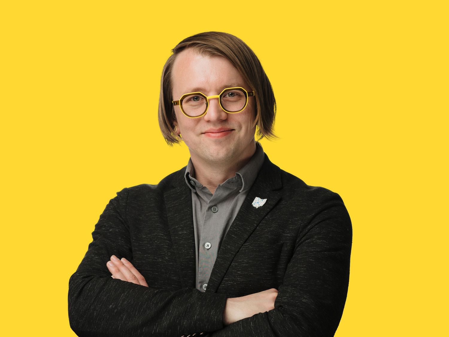 Person with short hair and yellow glasses wearing a dark blazer, arms crossed, against a bright yellow background.