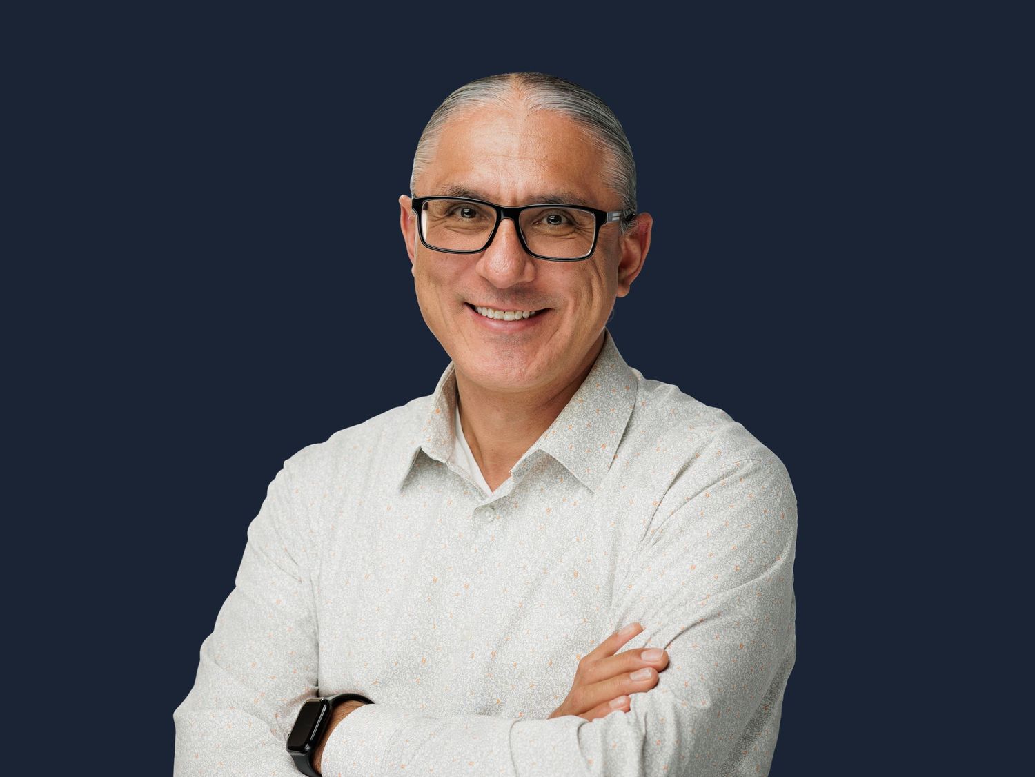 Smiling middle-aged man with glasses and crossed arms wearing a white shirt against a dark blue background.