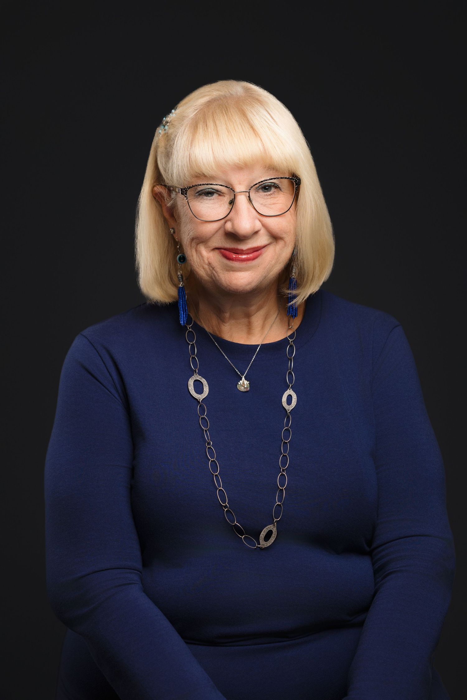 Professional portrait series of a person wearing glasses and a navy blue sweater against a dark background with silver necklace.