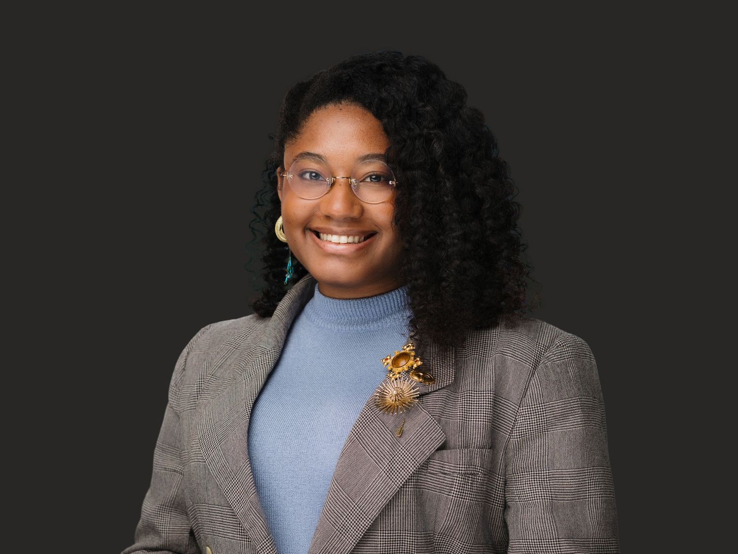 Young Black professional woman smiling in gray blazer with floral pin and blue turtleneck.
