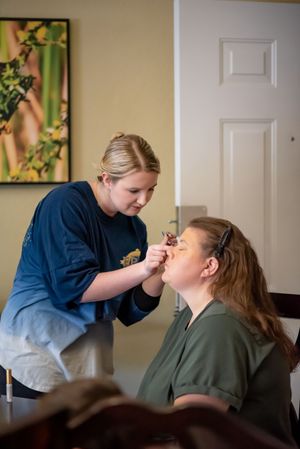 A makeup artist in a blue shirt applies makeup to a seated woman with auburn hair in a bright room.