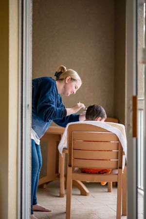 A woman feeding a young child sitting in a wooden chair at a dining table near a window.