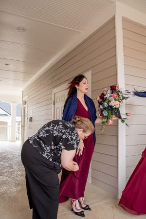 Women in formal attire prepare for a wedding on a covered porch with floral decorations.