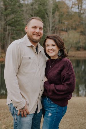 A couple stands together outdoors by a lake in fall, wearing casual sweaters and jeans.