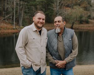 Two people in casual attire standing together and smiling by a serene lake with autumn trees in the background.