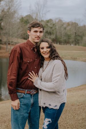 A couple poses together by a lake on an overcast day wearing casual fall attire in earth tones.