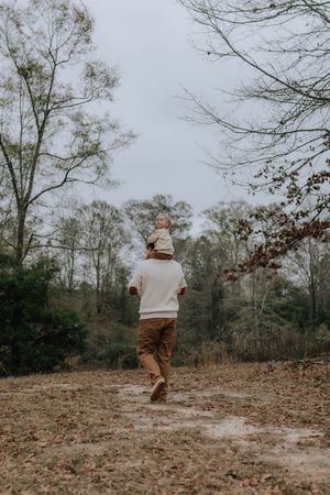 A person wearing a cream sweater and brown pants walks away on a leaf-covered dirt path through bare winter trees.