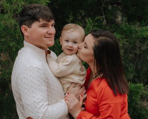 A family poses together outdoors, wearing a white sweater and red top, sharing a sweet moment with their baby.