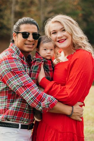 Family of three wearing coordinating red and plaid outfits for outdoor portrait.