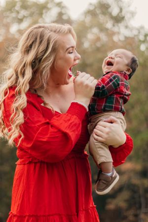 Playful moment between mother in red dress and laughing baby in plaid outfit.