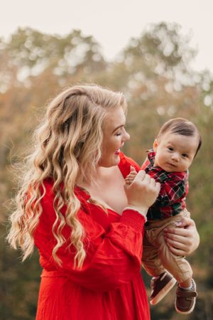 Mother in red dress holds baby wearing plaid outfit during autumn outdoor photoshoot.