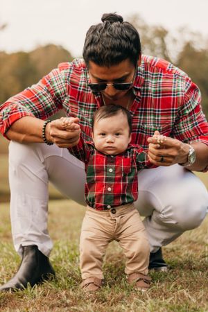 Parent in plaid shirt and sunglasses helps baby in matching outfit stand on grass.