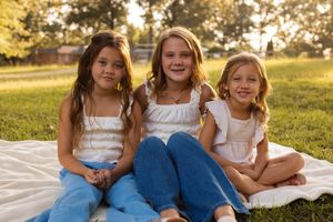 Three people in matching blue and white outfits sit together on a blanket in a sunny field during golden hour.