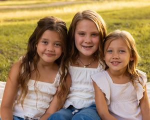 A group of children in white dresses sit together outdoors in golden sunlight during a photo session.