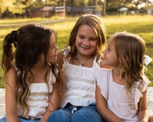 Three young friends in white tops share laughter and smiles while sitting together outdoors in warm sunlight.