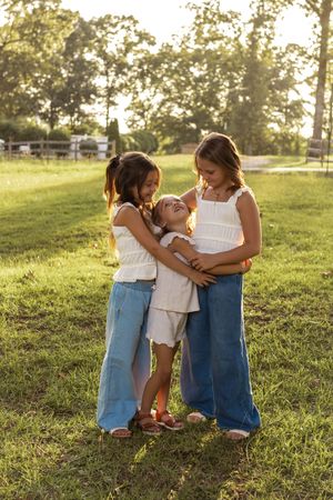 Three figures in casual summer attire share tender embraces on a sunlit grassy hillside during golden hour.