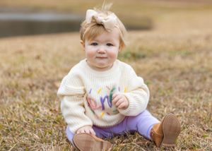A toddler wearing a cream sweater and purple pants sits on brown grass with decorative balloons.