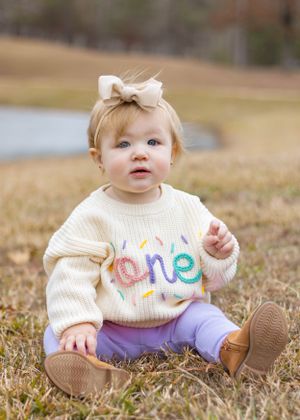 A toddler wearing a decorated cream sweater and purple pants sits on brown grass.