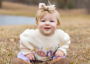 A toddler in a cream colored sweater smiles while sitting outdoors in autumn.