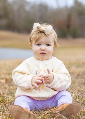 A toddler in a cream sweater and purple pants sits on dried grass outdoors.
