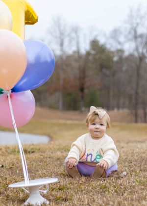 A toddler in cream sweater sits on grass with colorful balloons in the background.