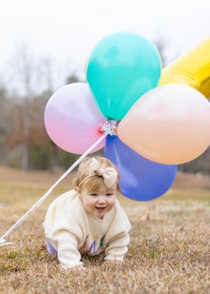 A toddler in cream sweater plays with colorful balloons on brown grass.