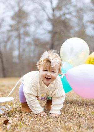 A joyful baby in cream sweater crawling on grass with colorful balloons in the background.