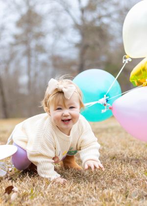 Baby in knit sweater laughing while playing with pink and blue balloons on brown grass.