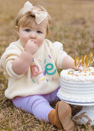 Baby in cream sweater with bow sits by white birthday cake with 'one' written in gold.