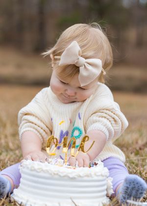 Small child in cream sweater reaching for gold 'one' cake topper on white birthday cake.