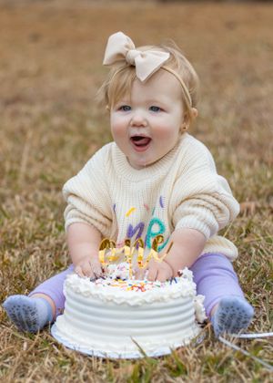 Baby with bow smiling excitedly at white birthday cake with gold decorations.