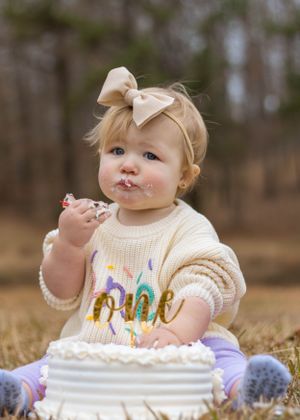 Baby with messy face enjoying first birthday cake while wearing cream sweater with colorful text.