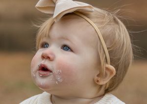 Close up of baby with cake frosting on face wearing cream bow and sweater.