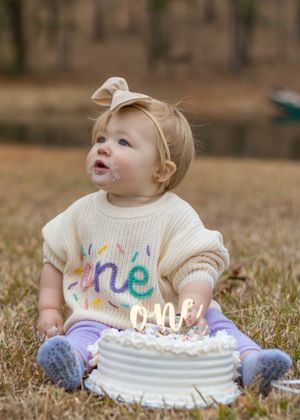 Baby in decorated sweater smiling at first birthday cake with gold 'one' topper.