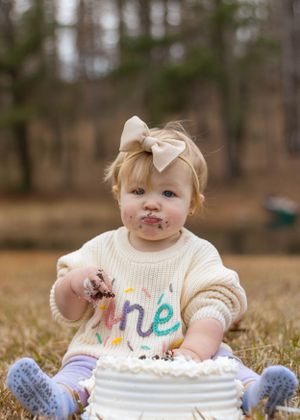 Baby with messy face reaching into white birthday cake while wearing cream sweater.
