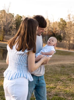 Couple in casual attire holding their baby outdoors near a lake at sunset.