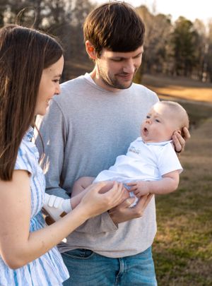 Parents in casual clothing tenderly hold their infant in a sunny outdoor setting.
