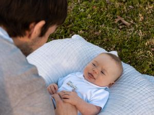 Baby in light blue outfit smiles while laying on quilted blanket outdoors.