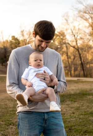 Parent in gray sweatshirt tenderly holding infant in white onesie outdoors during golden hour.