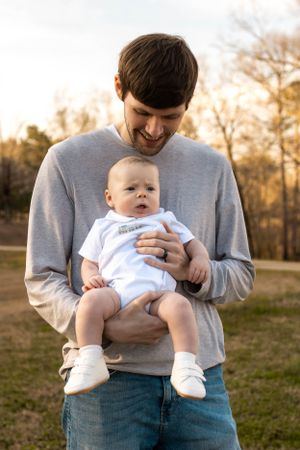 Baby in white onesie smiles while being held by adult in casual attire at sunset.