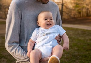 A smiling baby in a white onesie being held by someone wearing a gray long-sleeve shirt.