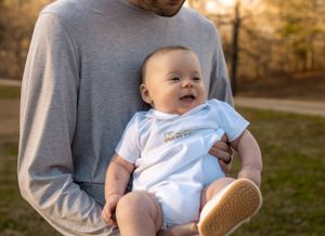Happy baby wearing white onesie being held against gray sweater outdoors.