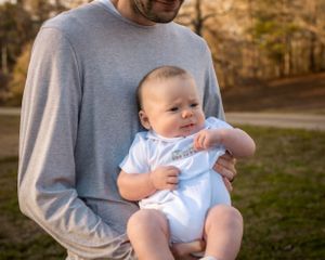 A baby wearing light blue clothing being held by someone in a gray sweater outdoors.