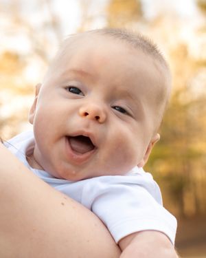 Close-up of smiling baby in white onesie with sunlit background.