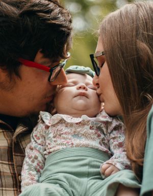 Parents sharing sweet kisses with baby between them in outdoor setting.